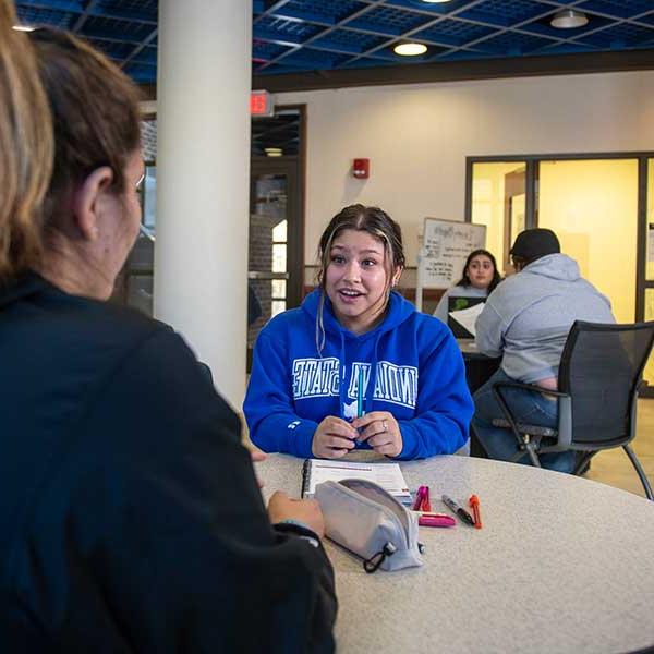 A student in a royal blue Indiana State sweatshirt is seated at a table in a commons area, speaking to another student who is mostly unseen. Two other students are visible at a table behind them.