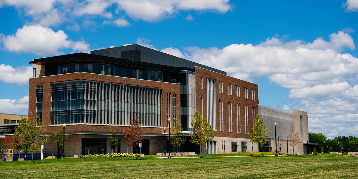 An exterior view of the College of Health and Human Services building, a brick building with numerous glass windows on the sides of the building. A green lawn is in front of the building.