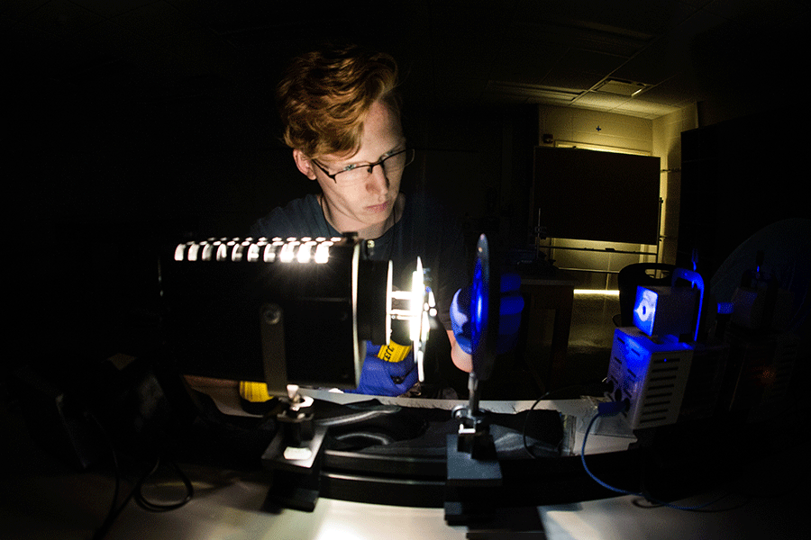 A male student with short red hair and glasses sits in a dark room. He is looking at physics lab equipment that emits an illuminating blue light.