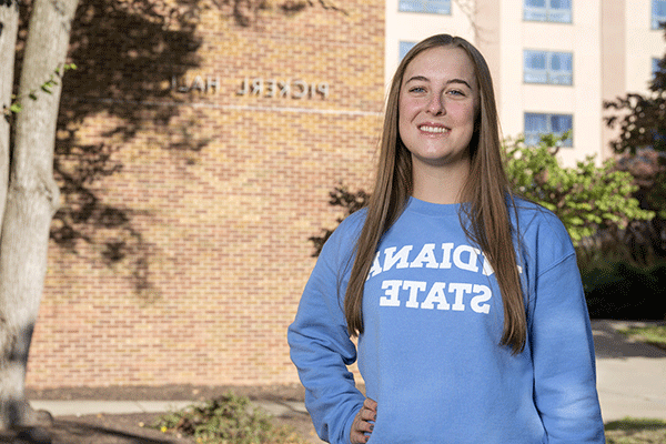 Portrait of a white female with long brown hair and wearing a light blue Indiana State long sleeved shirt looks at the camera with a brick building with “Pickerl Hall” on it in the background.