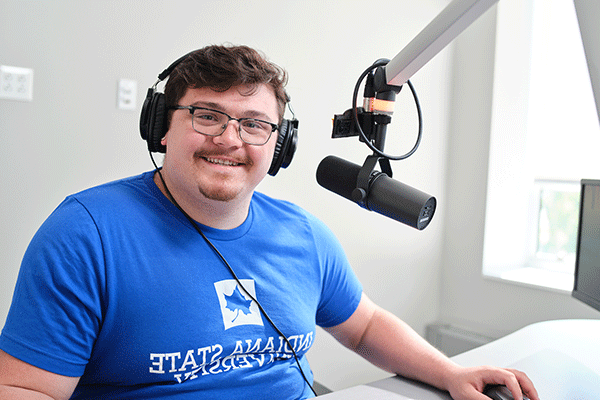 A white male student with short brown hair and glasses wearing a blue t-shirt with “Indiana State University” in white lettering. He looks and looking directly at the camera while wearing headphones and sitting in front of a radio microphone. 